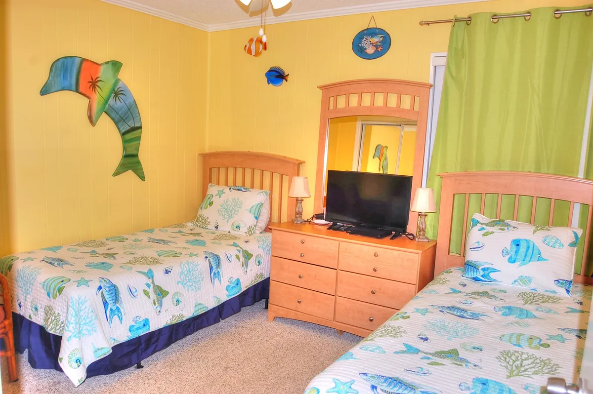 A playful second bedroom featuring two twin beds with aquatic-themed bedding, colorful dolphin wall art, a wooden dresser, and a television. The space feels inviting and is well-suited for families or groups, with ample storage and lively décor.