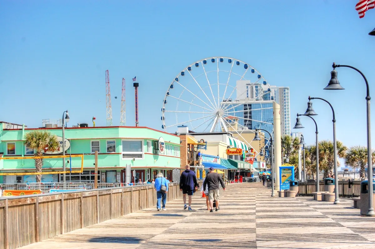 A vibrant exterior scene featuring the iconic Myrtle Beach Ferris wheel, a bustling boardwalk lined with colorful storefronts, and swaying palm trees. Strollers enjoy the lively atmosphere, and outdoor cafes invite guests to relax and take in the coastal energy.