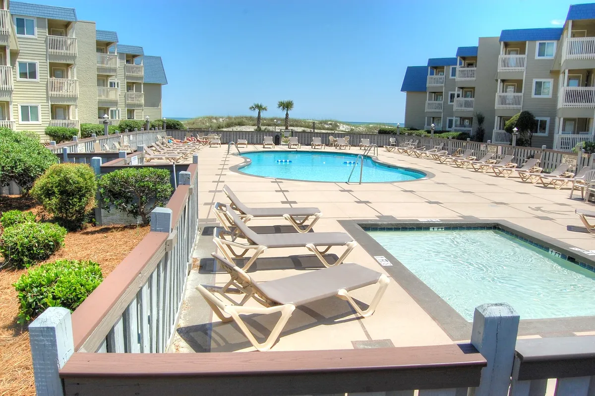 A large, sparkling swimming pool with an adjacent jacuzzi is surrounded by lounge chairs and lush landscaped greenery. In the background, low-rise buildings create a welcoming resort atmosphere, and a few guests are seen swimming, sunbathing, or relaxing by the pool.
