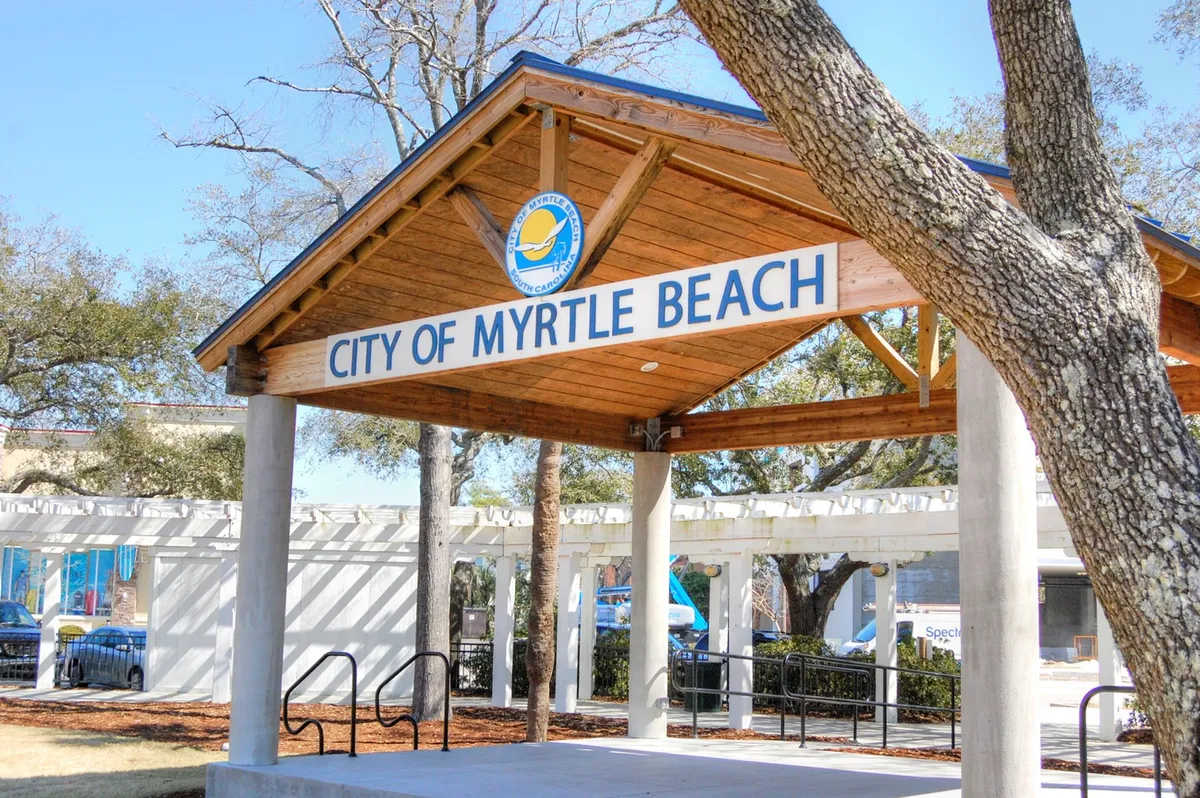 Features the 'CITY OF MYRTLE BEACH' sign on a wooden pavilion, accented by surrounding trees. The setting suggests a hub for community events, local gatherings, and informational resources for newcomers and seasonal guests.