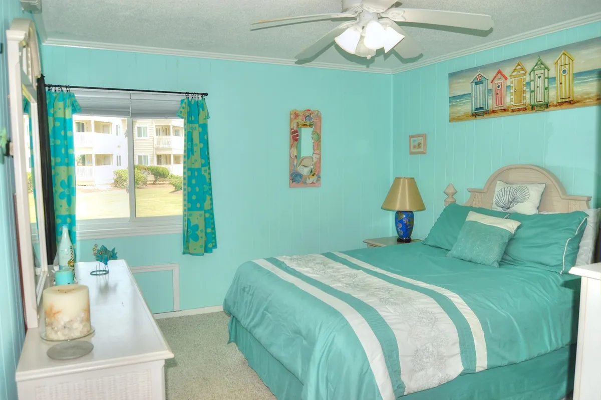 A bright, inviting bedroom with a comfortable bed, window letting in natural light, cheerful artwork, and a spacious dresser.