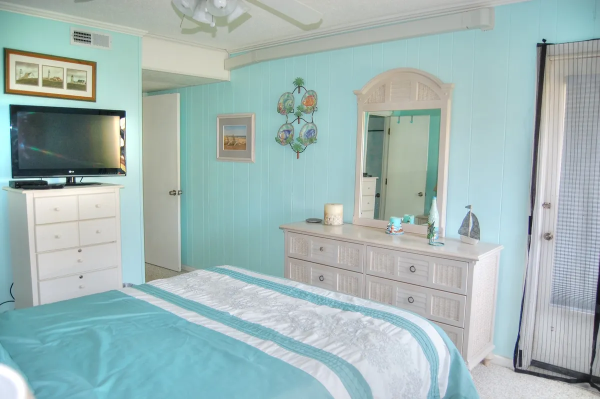 A tranquil bedroom with a blue accent wall, plush bed, dresser, mirror, and television. Natural light streams in, highlighting the room's calm ambiance and ample storage options.