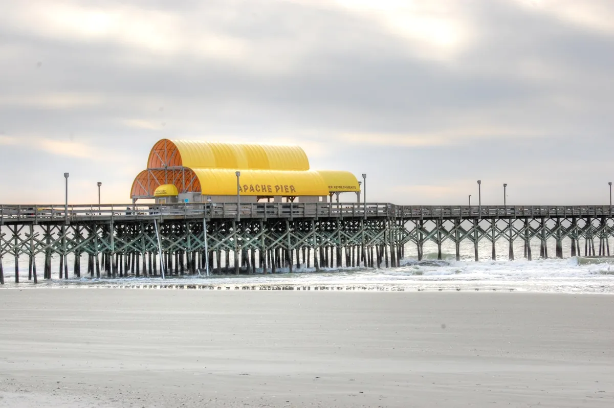 A tranquil view of the iconic Apache Pier stretches over gentle waves, supported by wooden beams, with people enjoying walks and fishing in the distance. The image conveys a sense of local character, relaxation, and connection to classic Myrtle Beach attractions.