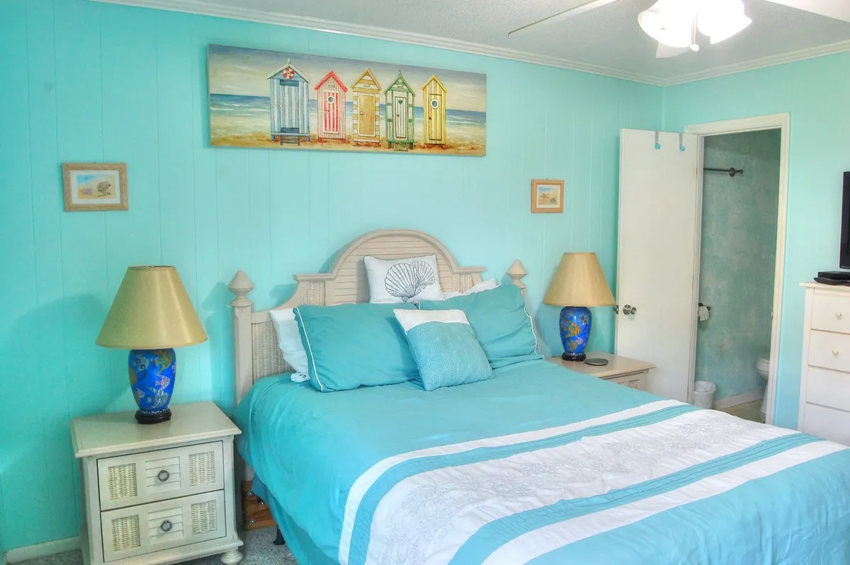 A tranquil bedroom with a bed dressed in turquoise bedding, complemented by beach-themed artwork and matching blue lamps. The serene color palette evokes an ocean-inspired atmosphere, and the soft lighting highlights the cozy linens and peaceful décor.