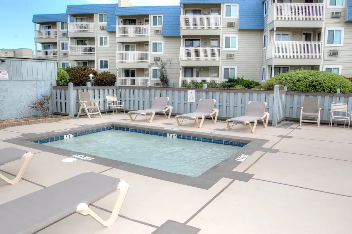 This image showcases a sparkling swimming pool surrounded by lounge chairs with the residential buildings of the complex visible in the background. The area is inviting and well-maintained, suggesting opportunities for relaxation and socializing with neighbors during sunny winter days.