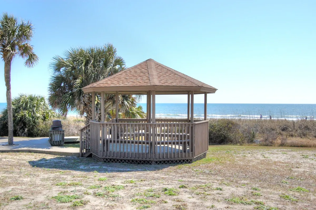 A serene oceanfront scene featuring a gazebo overlooking the beach and ocean, surrounded by palm trees. The area looks perfect for relaxing, picnicking, or enjoying quiet moments together with the sound of the waves in the background.