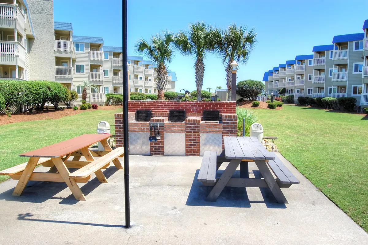 A vibrant grilling area featuring multiple picnic tables and standalone grills set on a lush green lawn, dotted with palm trees. Groups of people are barbecuing, dining, and socializing, capturing a relaxed, communal atmosphere.