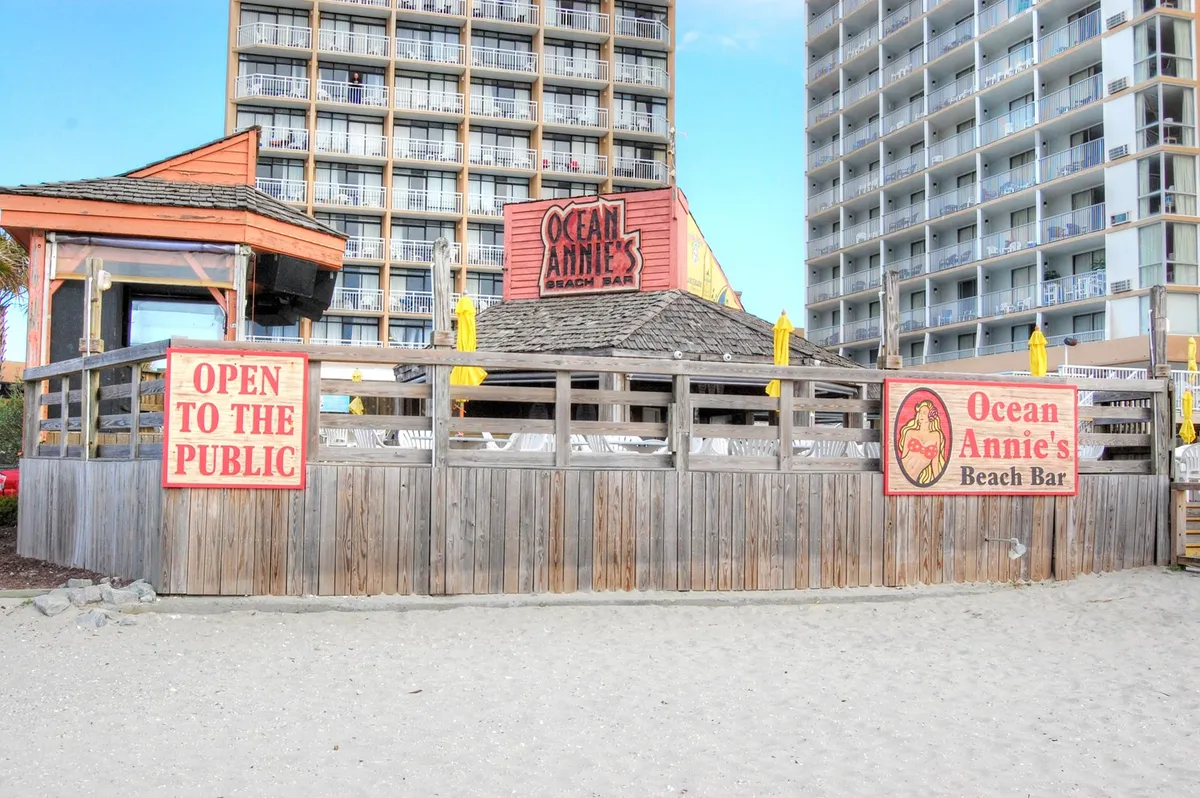 A vibrant beach bar with bright yellow umbrellas, wooden fencing, and a casual seating area directly overlooking the sand and sea. The inviting atmosphere suggests relaxed seaside dining and socializing as the sun sets.