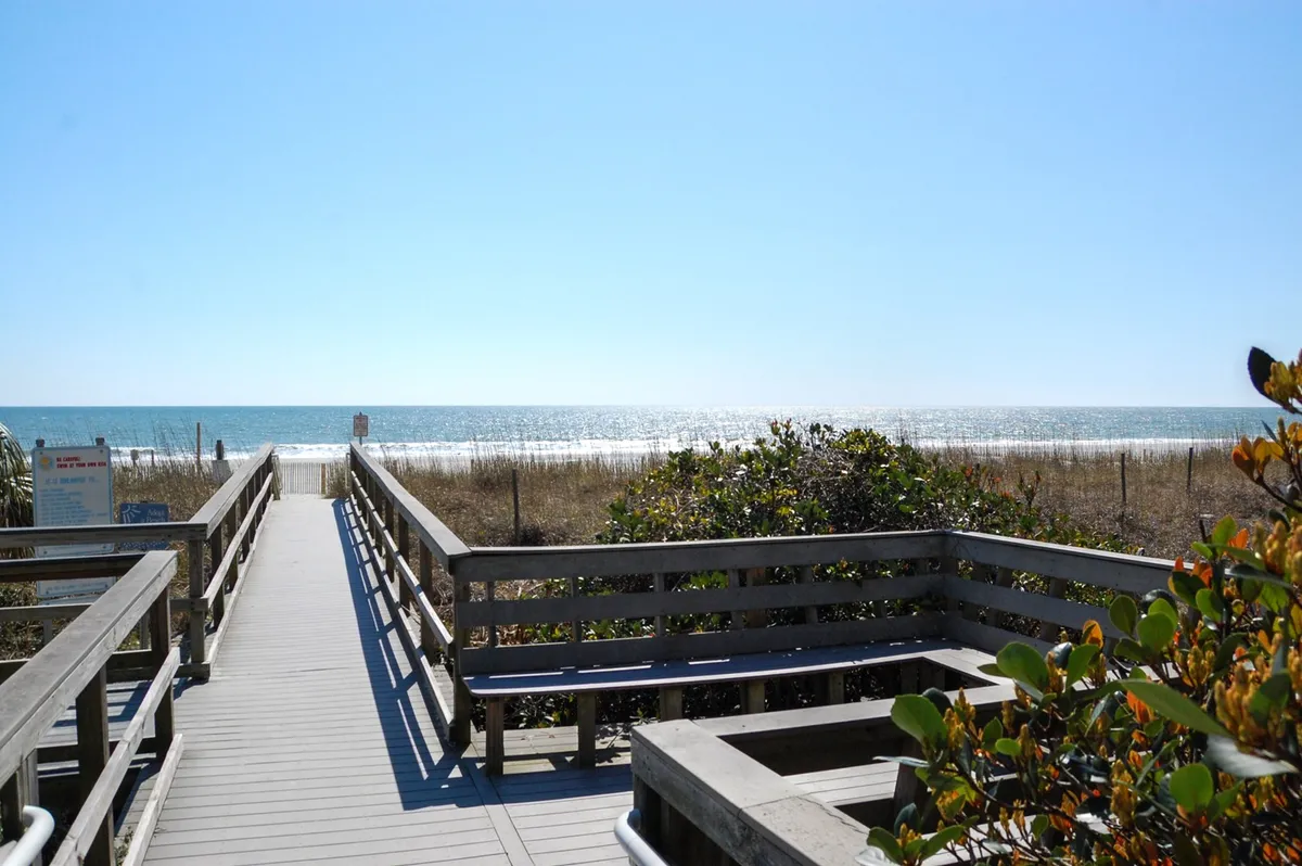 Depicts a scenic boardwalk stretching over the dunes toward the ocean, with natural vegetation and sweeping water views. The tranquil setting conveys the opportunity for relaxing walks, beach exploration, and enjoying sunsets along the quiet shore.