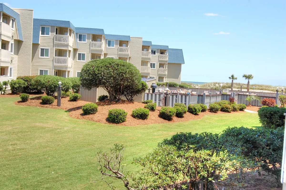 A serene landscaped lawn leads to a pathway that extends directly toward the beach, with balconies of the condo visible in the background. The scene is calm and inviting, illustrating direct access from the property to the beach and outdoor spaces for relaxation.
