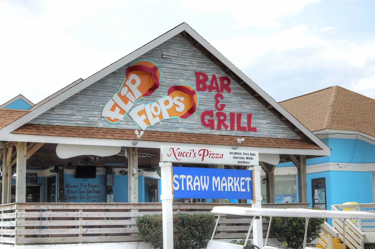 A cluster of welcoming storefronts with signage for Flip Flops Bar & Grill, Nucci's Pizza, and the Straw Market. Outdoor seating encourages socializing and al fresco dining, while the setting sun adds a relaxed, coastal vibe.